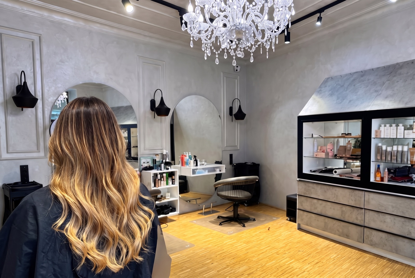 Balayage Wien – Kundin mit goldblondem Balayage-Farbverlauf im Salon mit Bogenspiegeln und Kristallluster